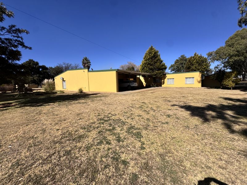 Rear view of the property featuring a yellow facade, integrated carport, and expansive surrounding garden area.
