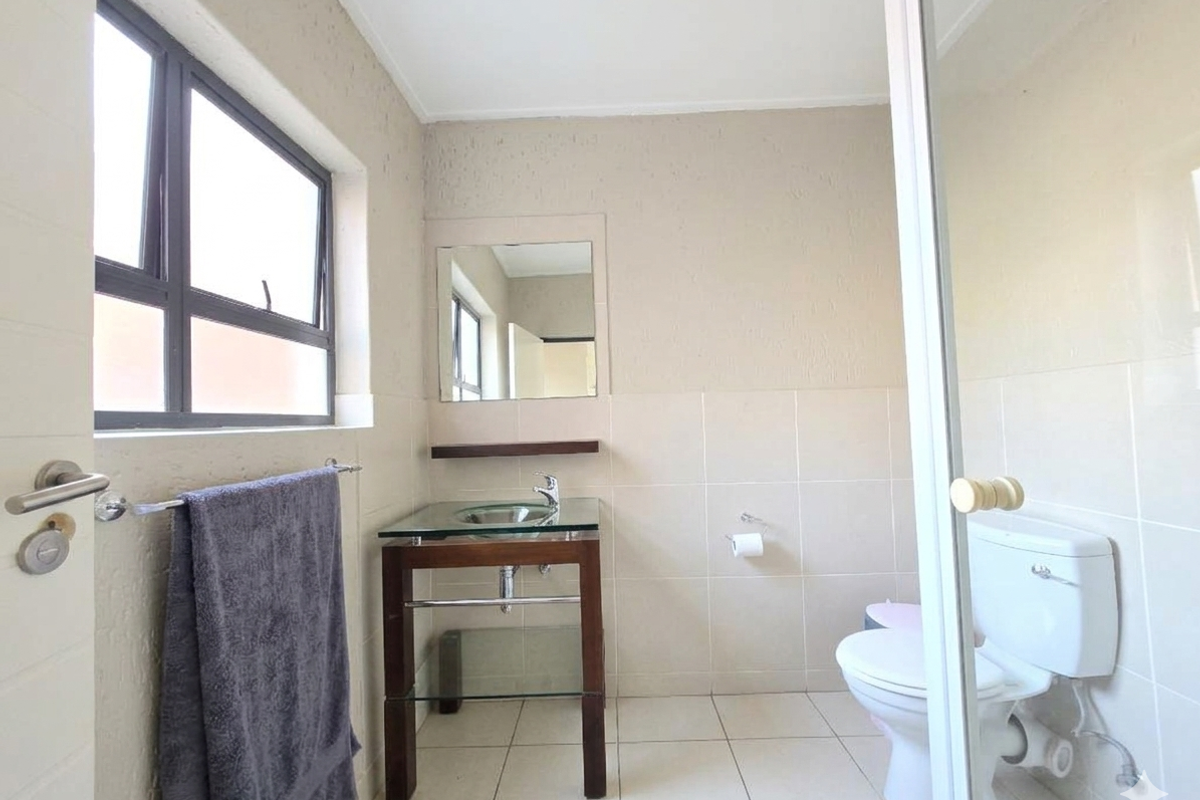 A functional bathroom featuring a glass vessel sink, wall-mounted mirror, toilet, and neutral tiled finishes with natural light from the window.