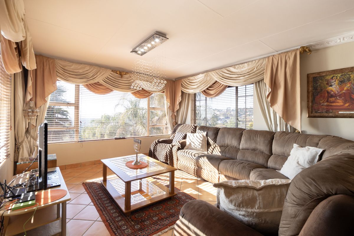 A sunlit living room featuring large windows with venetian blinds, tiled flooring, and a central ceiling light fixture.