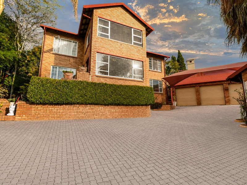 Front view of a multi-story brick townhouse featuring a spacious paved driveway, triple garage with roll-up doors, and established garden landscaping.