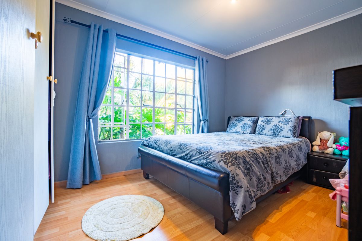 A well-lit bedroom featuring laminate flooring, a built-in cupboard, and a large window with security bars overlooking greenery.