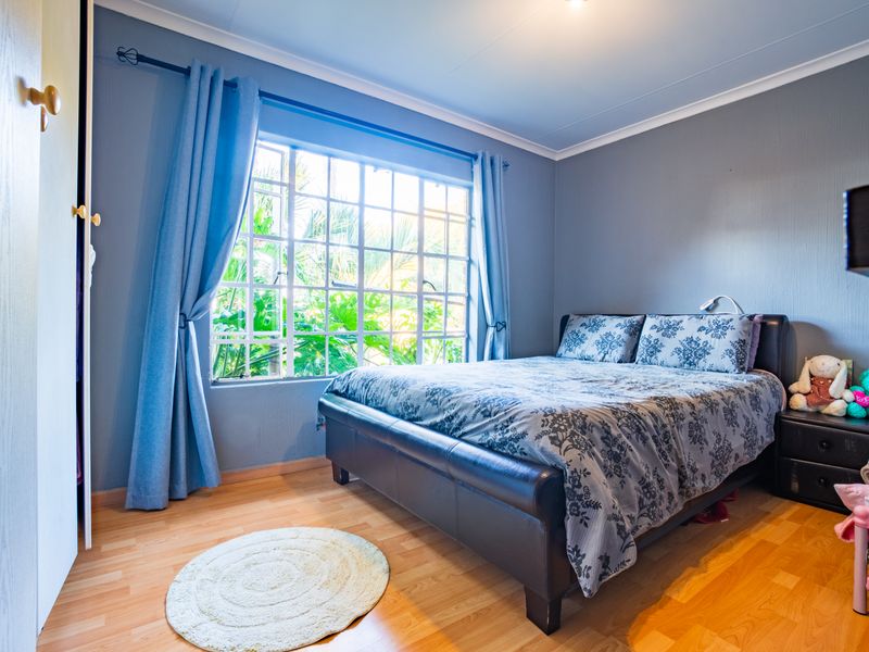 A well-lit bedroom featuring laminate flooring, a built-in cupboard, and a large window with security bars overlooking greenery.