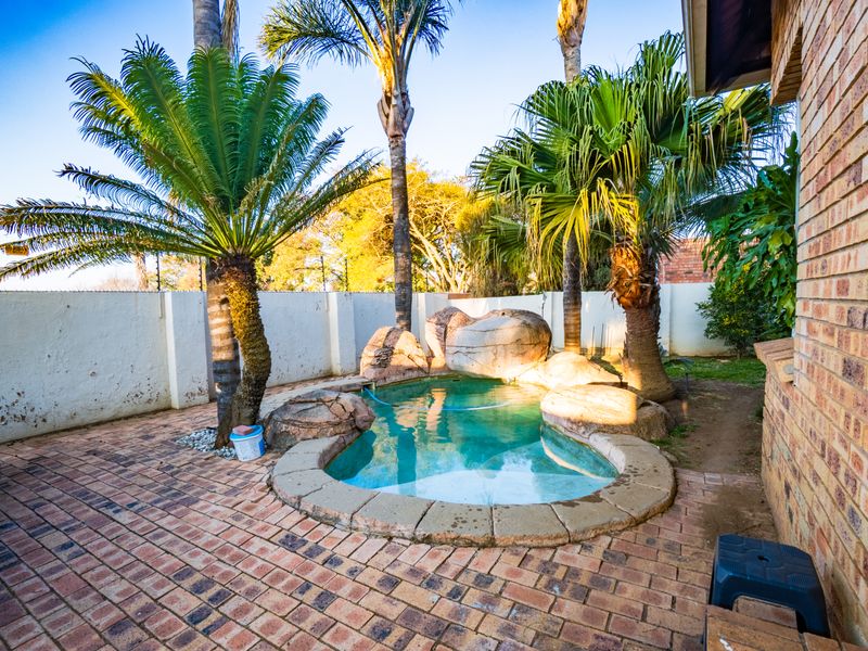 A compact backyard pool area featuring a rock-lined swimming pool, brick paving, and established palm trees within a private walled garden.