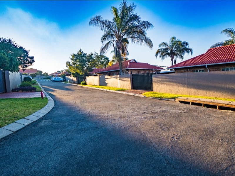 A view of the residential street within the townhouse complex, featuring paved roads, perimeter walls, and established landscaping.