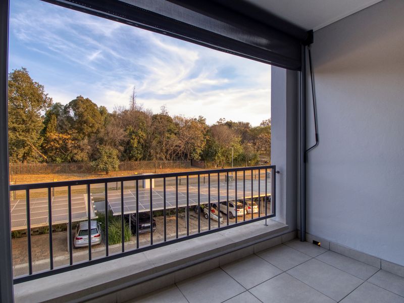 A covered balcony featuring tiled flooring, a metal balustrade, and a retractable outdoor blind, overlooking the parking area and surrounding trees.