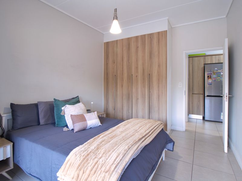 A well-maintained bedroom featuring modern wood-finish built-in cupboards, neutral tiled flooring, and a clean, contemporary aesthetic with direct access to the kitchen area.