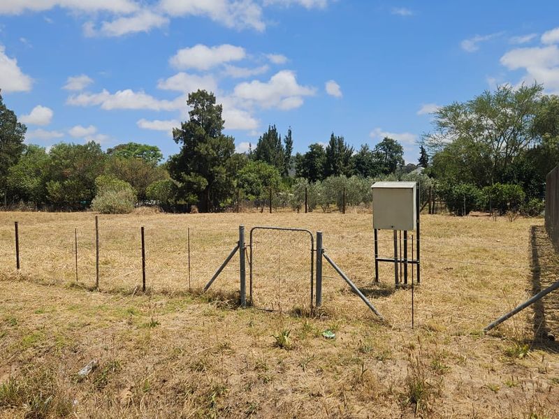 A view of a vacant residential plot featuring wire fencing, a small metal gate, and an installed electrical utility box.