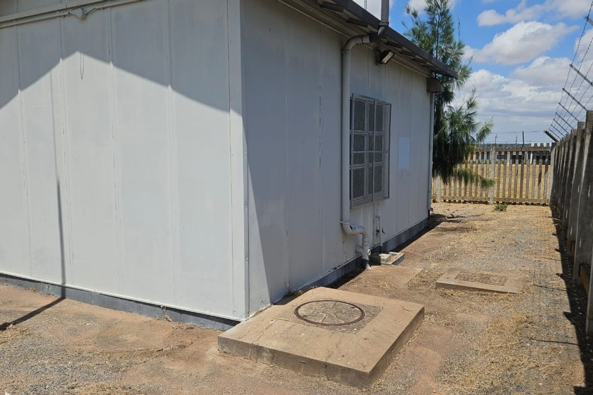 Rear view of a prefabricated structure featuring security fencing, external plumbing, and concrete utility access points on a dry, unlandscaped plot.