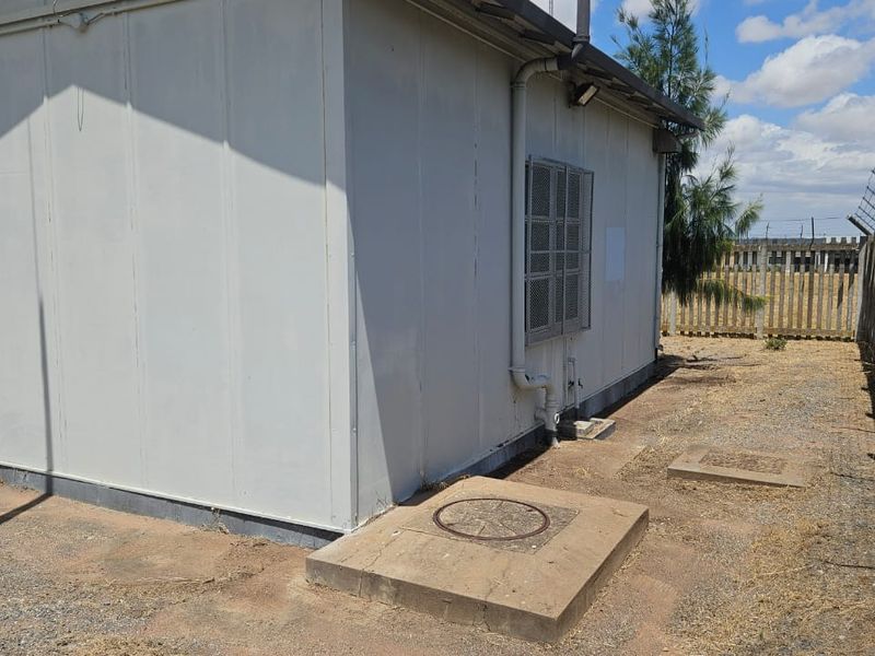 Rear view of a prefabricated structure featuring security fencing, external plumbing, and concrete utility access points on a dry, unlandscaped plot.