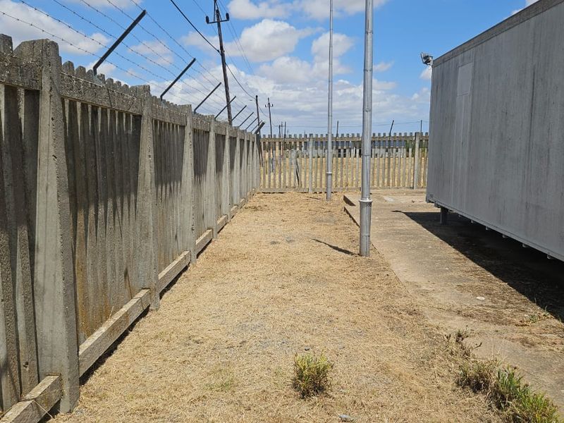 A side yard area featuring concrete palisade fencing with barbed wire, a concrete slab, and a metal utility structure.