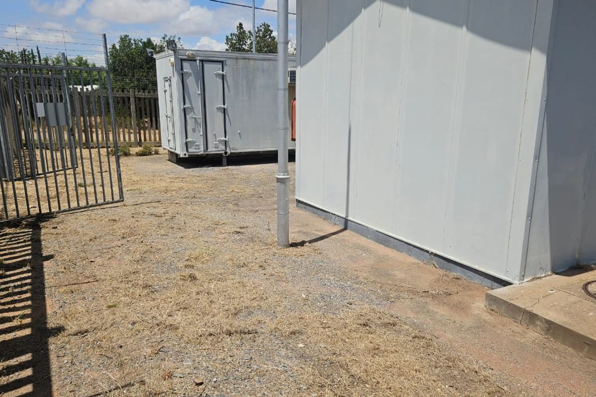 A view of the rear exterior area featuring a prefabricated structure, gravel ground cover, and metal security fencing with barbed wire.