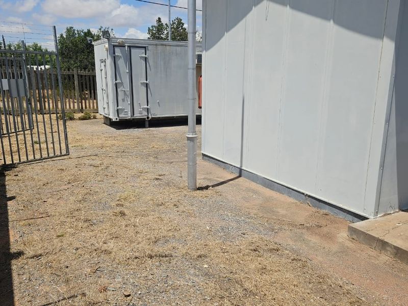 A view of the rear exterior area featuring a prefabricated structure, gravel ground cover, and metal security fencing with barbed wire.