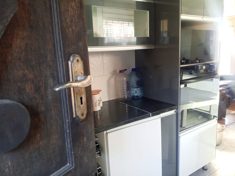 A compact kitchen area featuring modern high-gloss cabinetry, built-in oven and microwave, and granite countertops, viewed through a wooden doorway.