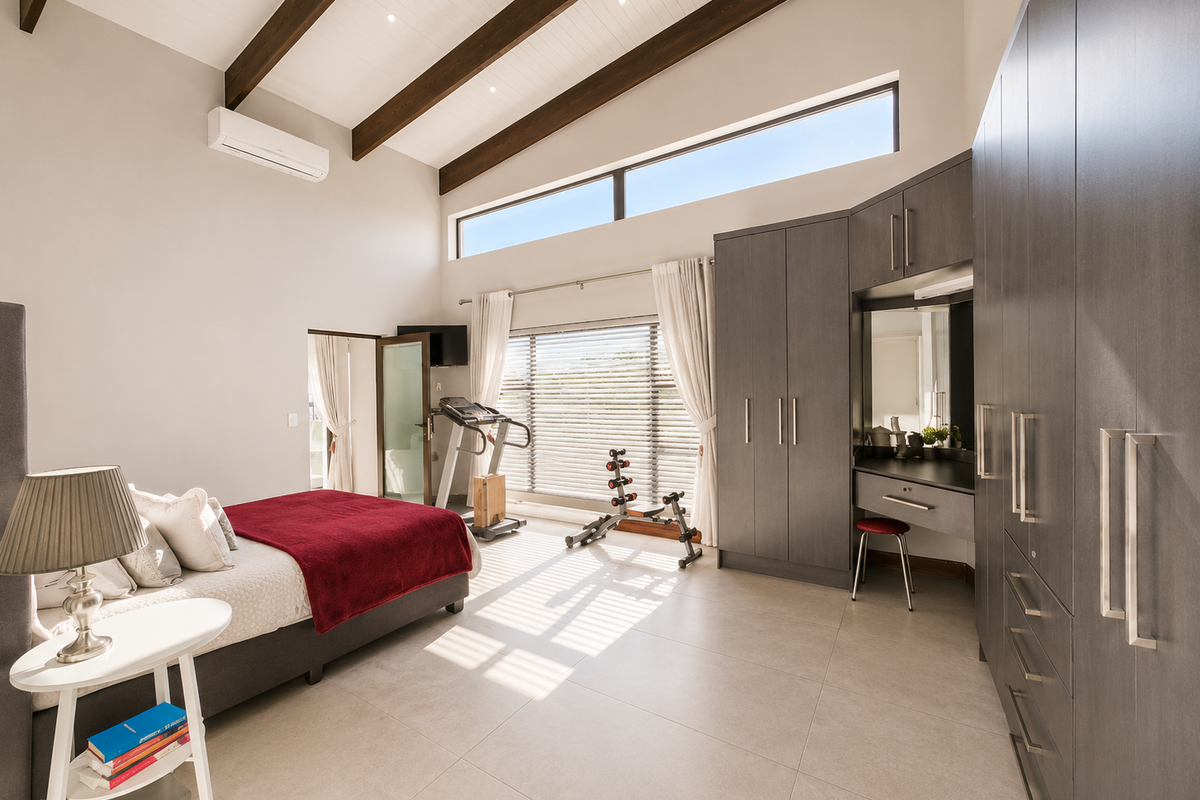 Spacious bedroom featuring vaulted ceilings with exposed timber beams, extensive built-in cabinetry with integrated vanity, tiled flooring, and large windows providing ample natural light.