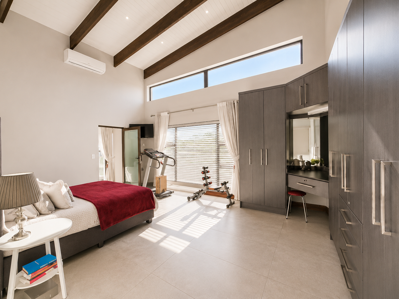 Spacious bedroom featuring vaulted ceilings with exposed timber beams, extensive built-in cabinetry with integrated vanity, tiled flooring, and large windows providing ample natural light.
