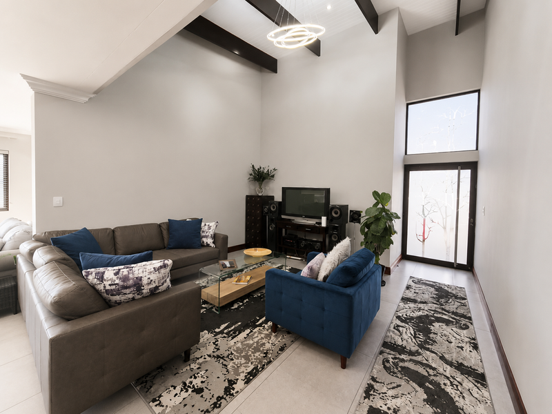 Spacious living room featuring high vaulted ceilings with exposed timber beams, large windows for natural light, and neutral tiled flooring.