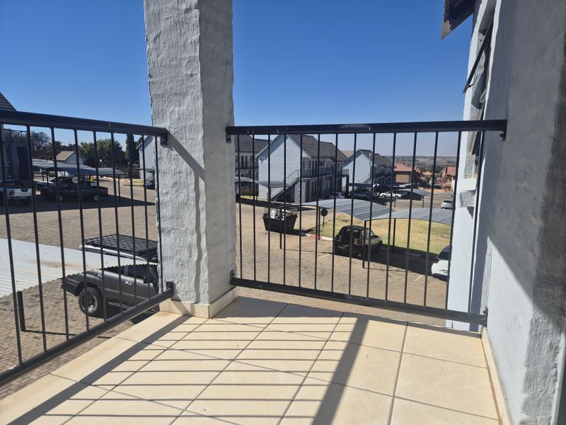 A private balcony featuring tiled flooring and a metal balustrade, offering an elevated view over the apartment complex parking area.