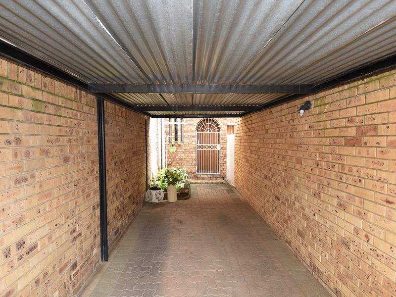 A covered carport area featuring brick walls, a corrugated metal roof, and paved flooring leading to a secure gated entrance.