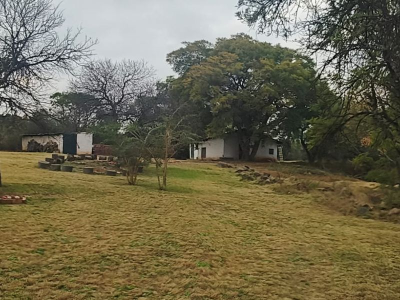 A wide view of a rural farm garden featuring mature trees, natural rock landscaping, and a small outbuilding in the distance.