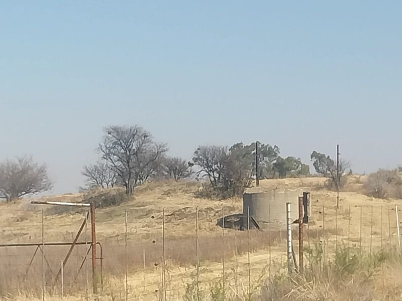 A view of the small holding grounds featuring a concrete water reservoir, perimeter wire fencing, and a metal gate on a dry landscape.