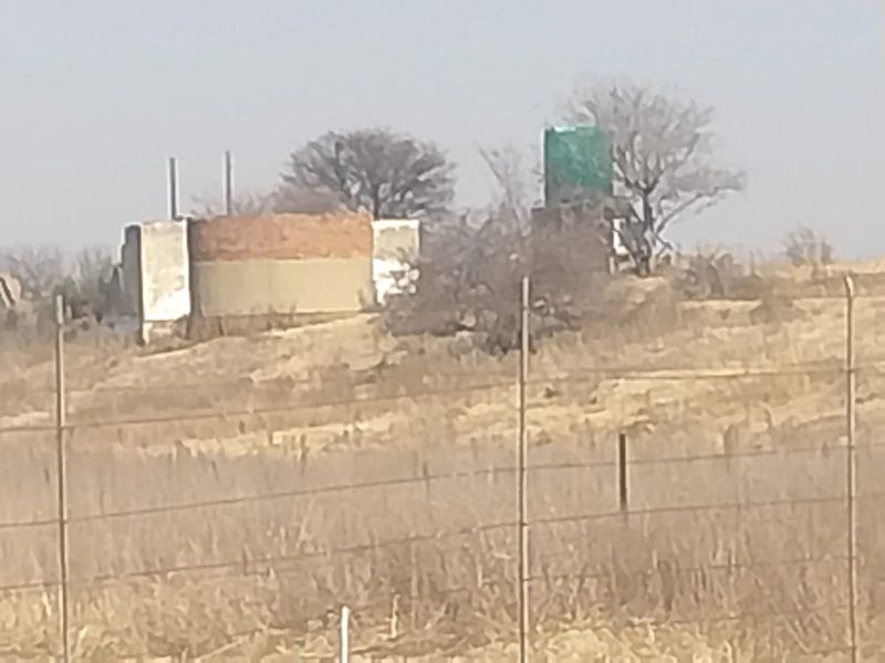 Distant view of a small holding property featuring an unfinished masonry structure and a water storage tank, enclosed by perimeter fencing.