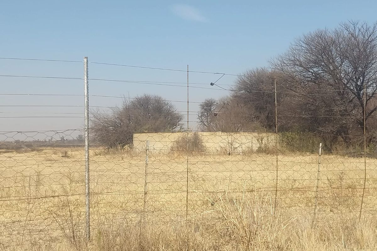 A view of a rural small holding property featuring wire mesh fencing and a weathered concrete boundary wall set against dry, open land.
