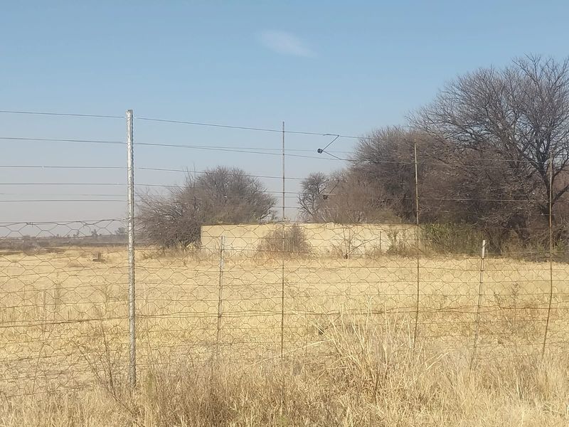 A view of a rural small holding property featuring wire mesh fencing and a weathered concrete boundary wall set against dry, open land.