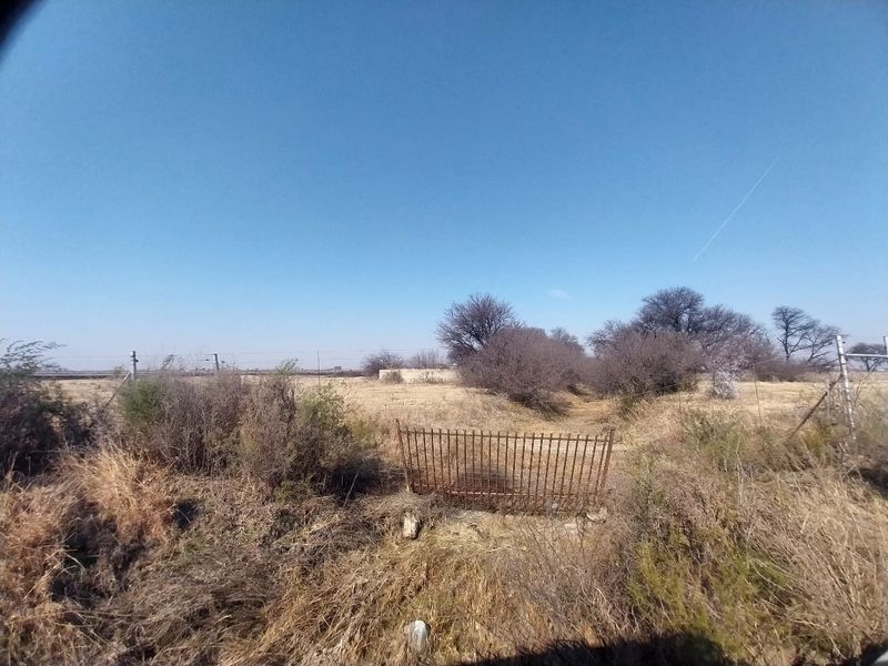 An expansive, undeveloped plot of land featuring dry vegetation, wire perimeter fencing, and a weathered metal gate entrance.