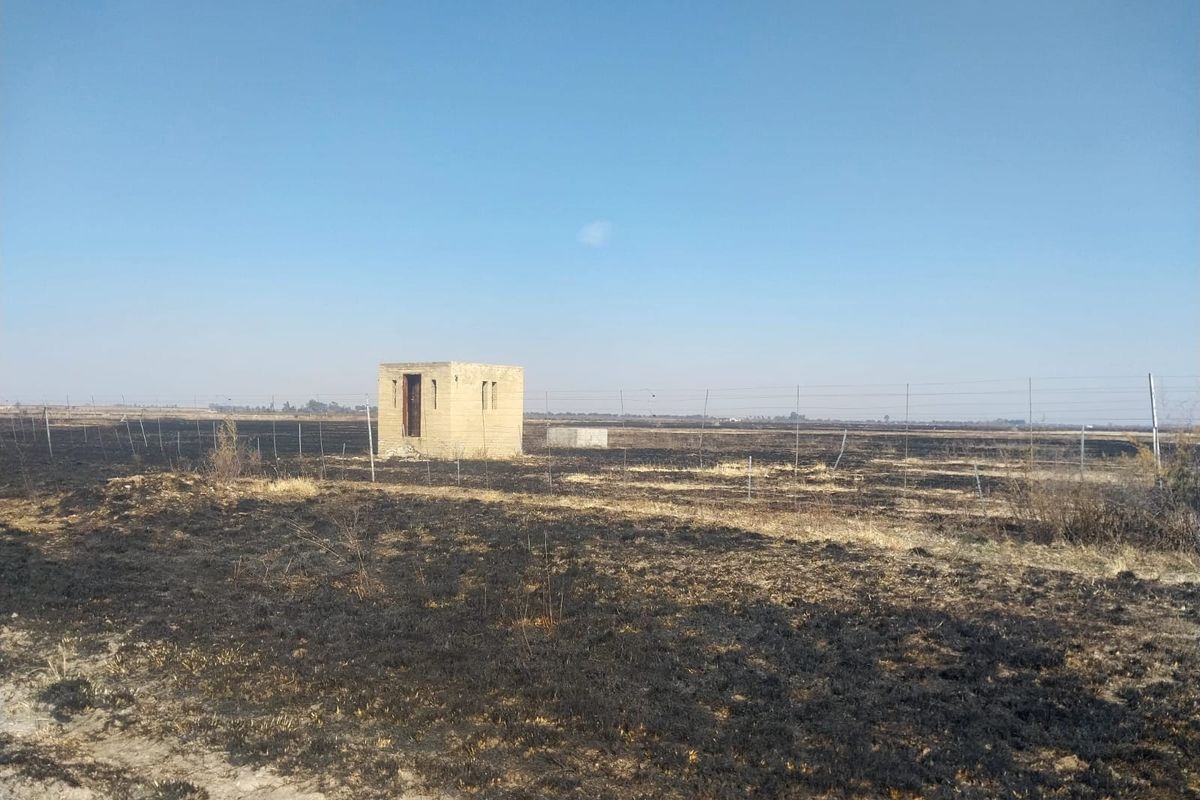 A view of a small brick utility structure situated on a large, open plot of land showing signs of recent fire damage.