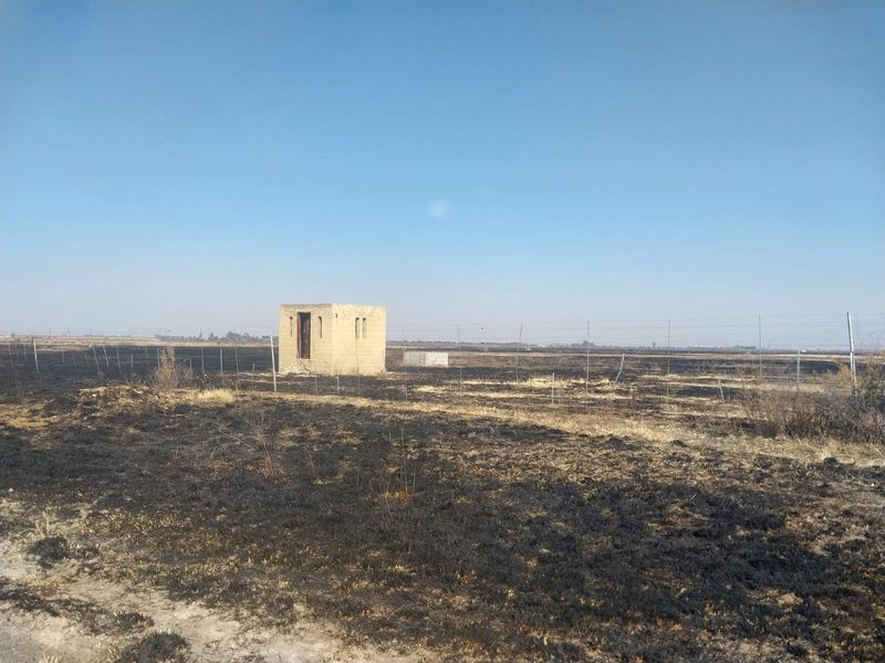 A view of a small brick utility structure situated on a large, open plot of land showing signs of recent fire damage.
