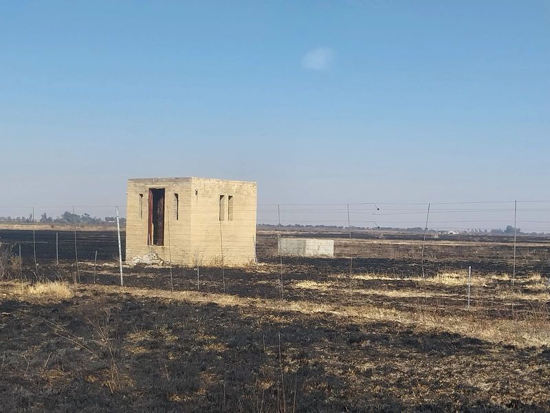 A small concrete outbuilding situated on a large, open plot of land with wire fencing and evidence of recent fire damage.