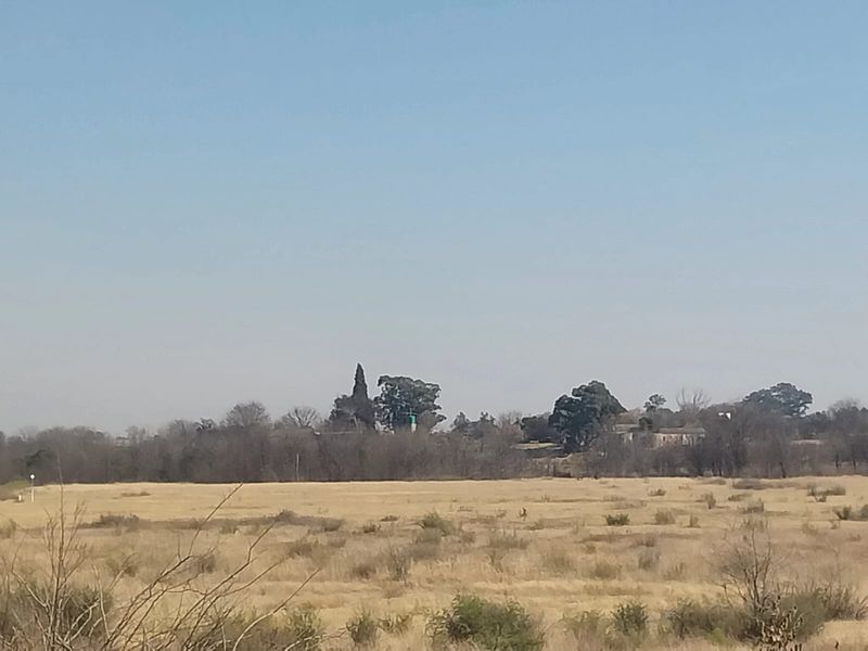 A wide landscape view of the small holding property featuring open fields and distant mature trees under a clear sky.