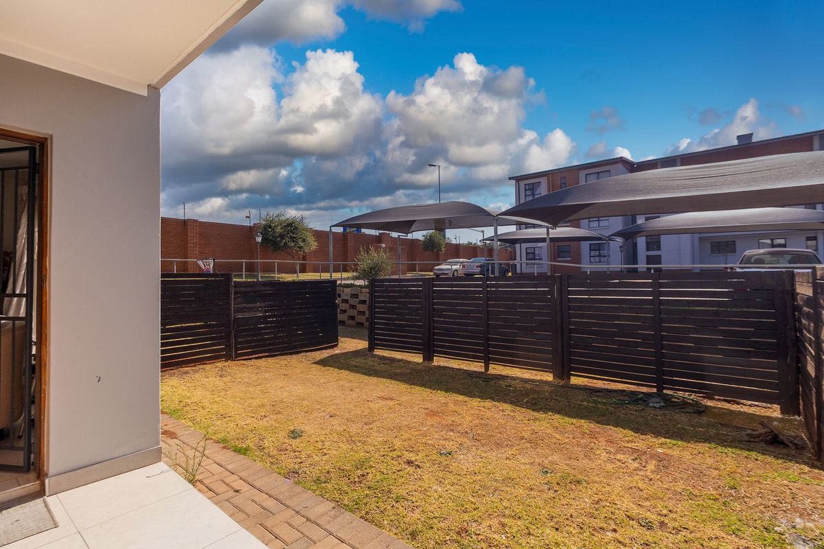A private rear garden area featuring a paved patio, timber fencing, and a view of the complex parking area with shade cloth carports.