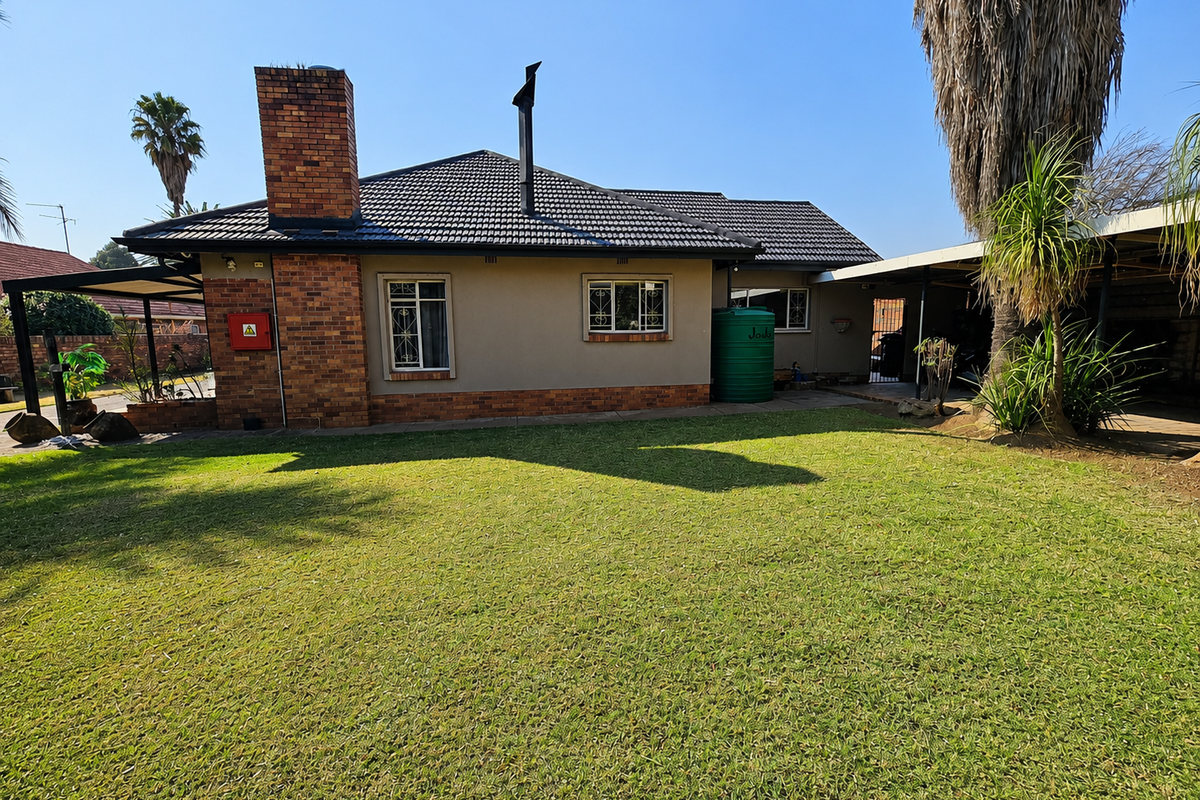 Rear view of the house featuring a brick chimney, tiled roof, water tank, and a spacious lawn area with an adjacent carport.