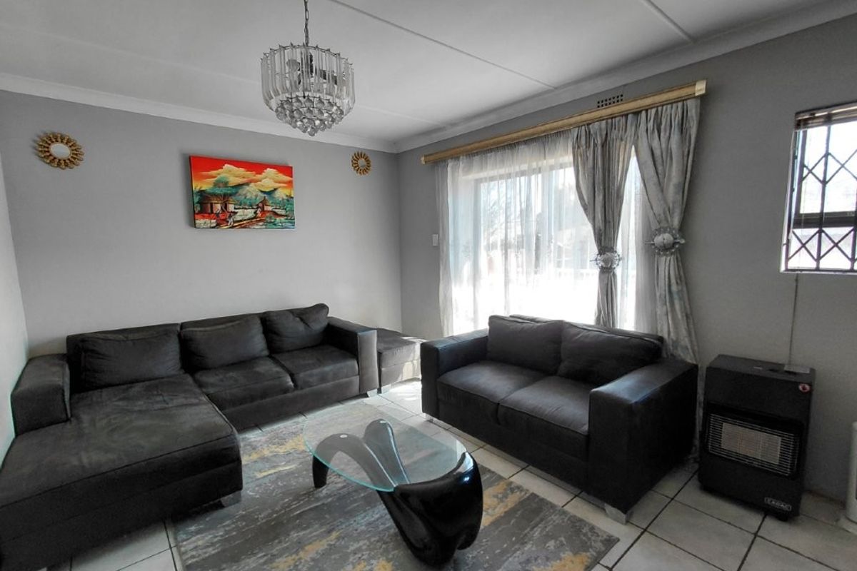 A well-maintained living room featuring tiled flooring, a decorative crystal chandelier, and a large window fitted with a security gate.