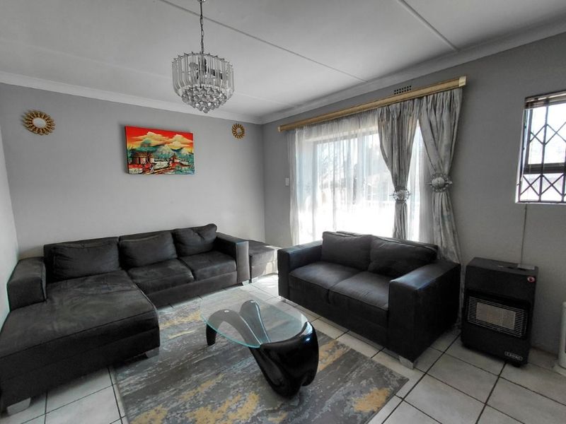 A well-maintained living room featuring tiled flooring, a decorative crystal chandelier, and a large window fitted with a security gate.