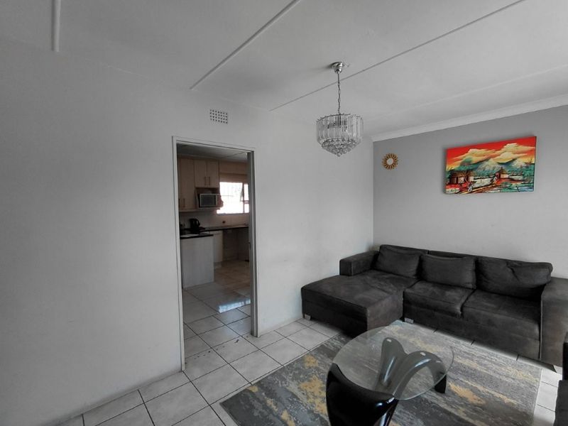 A living room featuring tiled flooring and a central light fixture, with an open doorway leading into the adjacent kitchen area.