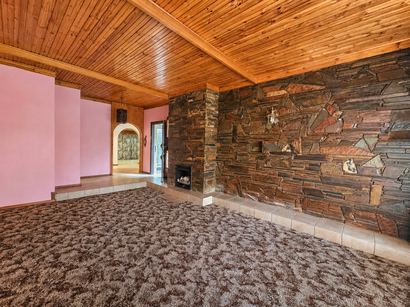 Spacious living room featuring a prominent stone fireplace, timber-clad ceiling, and tiled hearth, offering a unique architectural foundation for renovation.