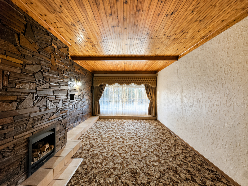 Spacious living room featuring a prominent stone fireplace, tiled hearth, and timber-clad ceiling, offering a blank canvas for modern renovation.