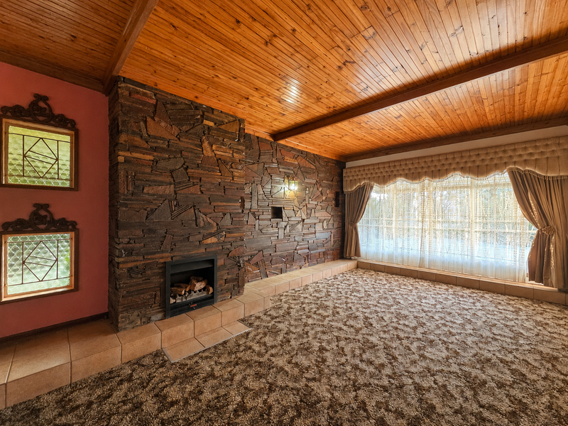 Spacious living room featuring a prominent stone fireplace, timber-clad ceiling with exposed beams, and decorative windows, offering a classic, dated aesthetic ready for modernization.