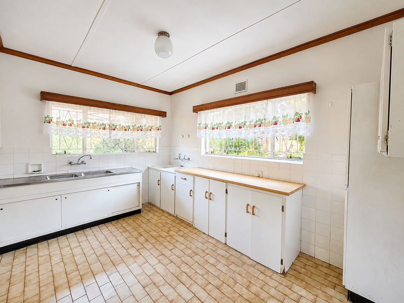 A spacious, dated kitchen featuring original white cabinetry, laminate countertops, tiled splashbacks, and ceramic floor tiles, offering significant potential for a modern renovation.