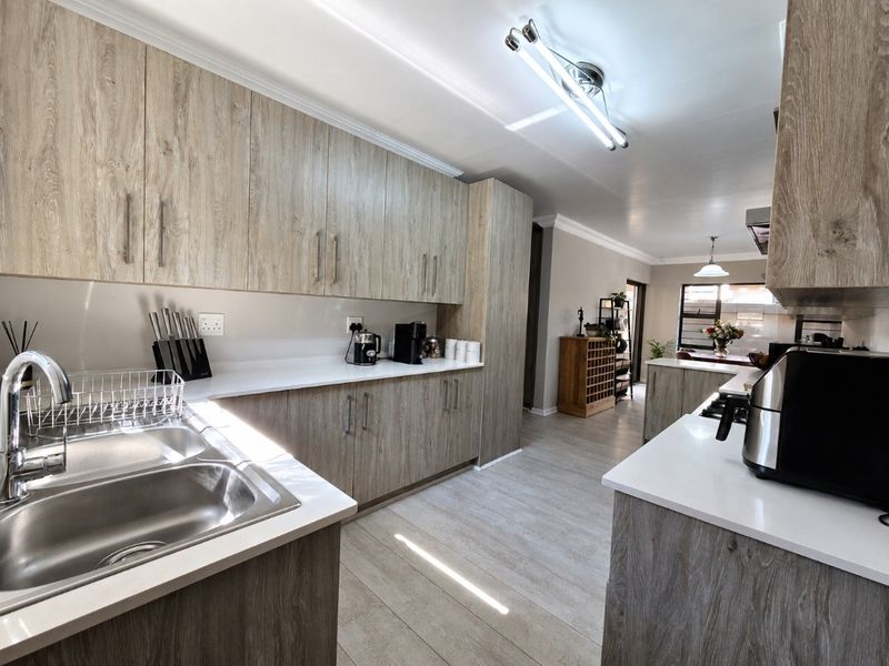 Modern kitchen featuring light wood-grain cabinetry, white quartz countertops, double stainless steel sink, and integrated hob with extractor fan, flowing into an open-plan living area.