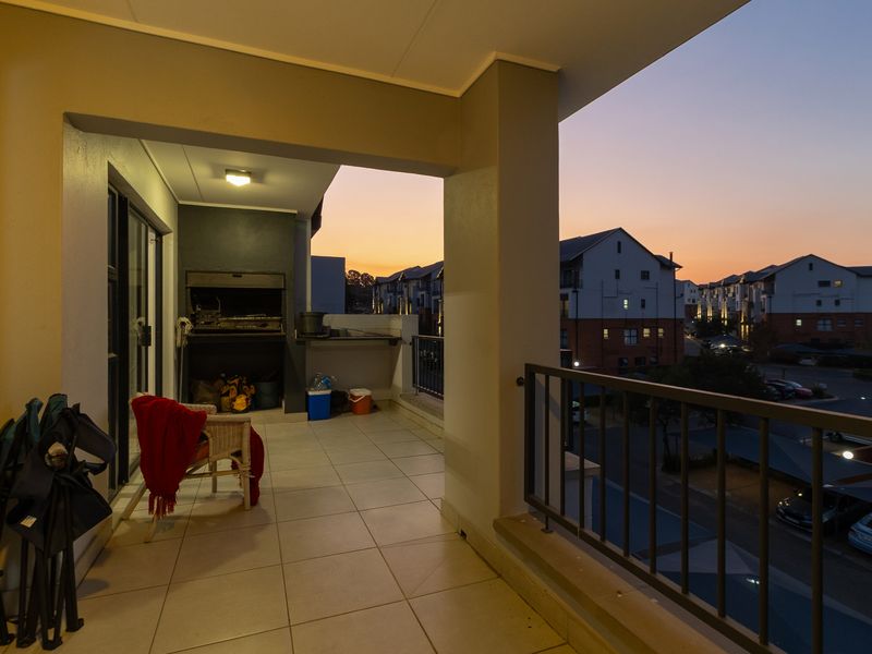 Covered balcony featuring a built-in braai, tiled flooring, and metal balustrade, offering an elevated view of the surrounding apartment complex at sunset.