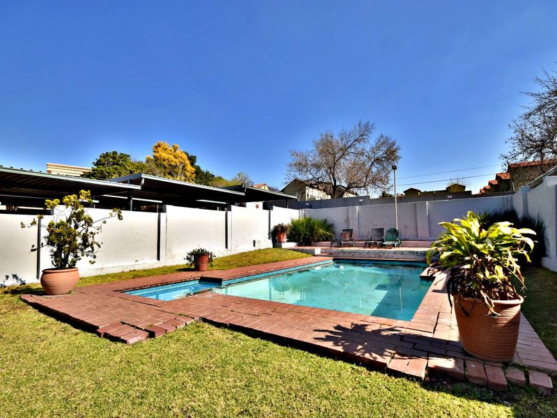 Outdoor pool area featuring a swimming pool with brick coping, surrounded by a lawn and perimeter wall, with an adjacent carport structure.
