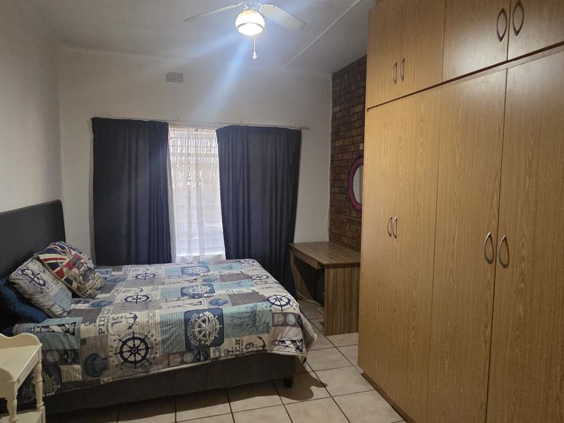 A bedroom featuring built-in wooden cupboards, an exposed brick feature wall, tiled flooring, and a ceiling fan.