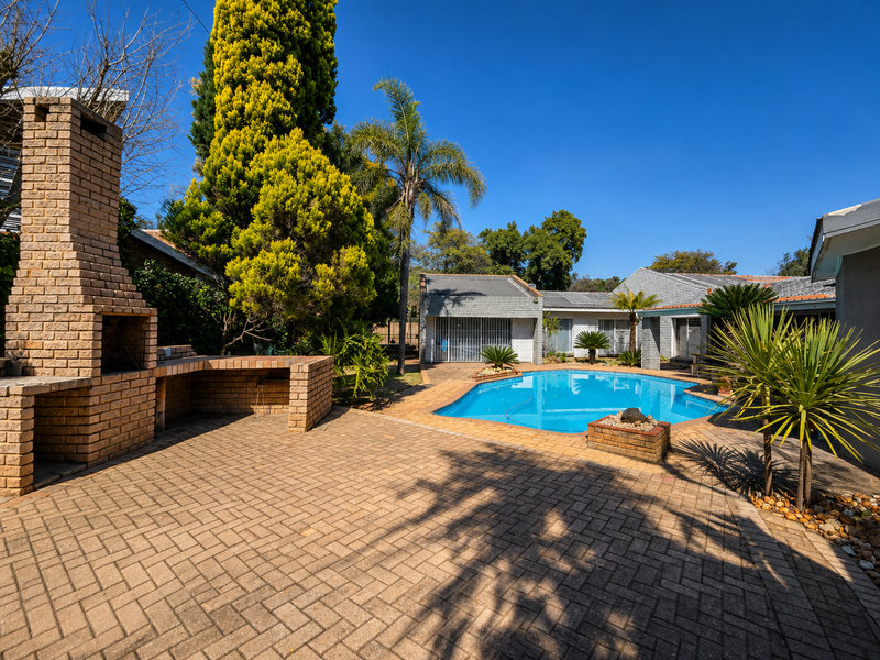 Outdoor entertainment area featuring a paved patio, built-in brick braai, and a swimming pool set within a landscaped garden.