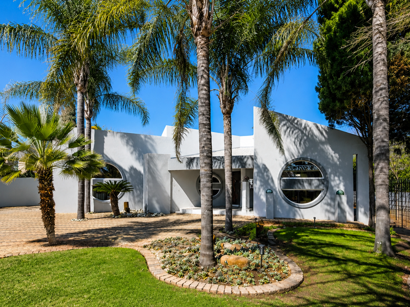 A well-maintained front facade featuring unique circular windows, a paved driveway, and established palm trees in a landscaped garden setting.