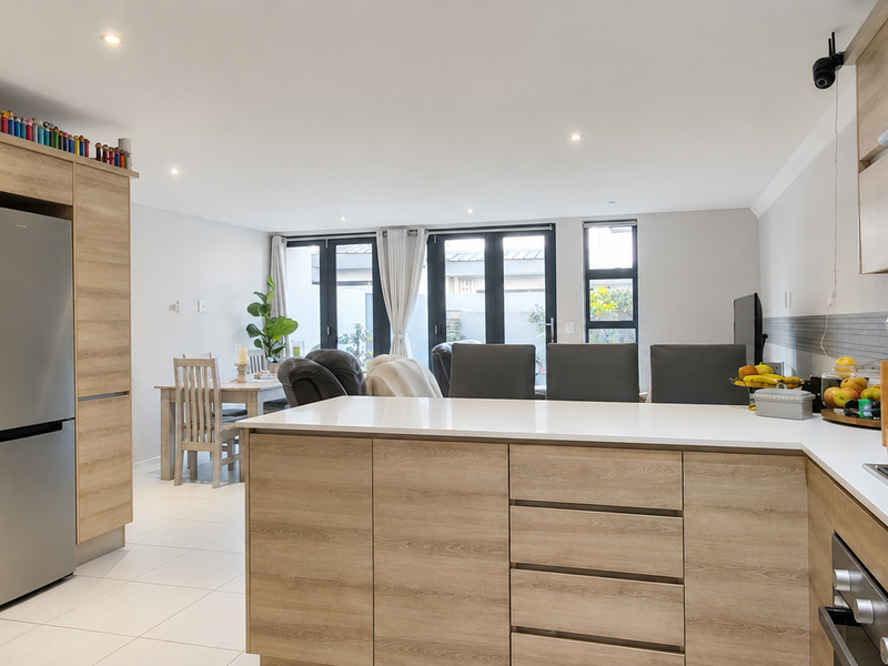 Modern open-plan kitchen and living area featuring light wood cabinetry, stone countertops, integrated appliances, and direct access to the outdoor space through sliding doors.