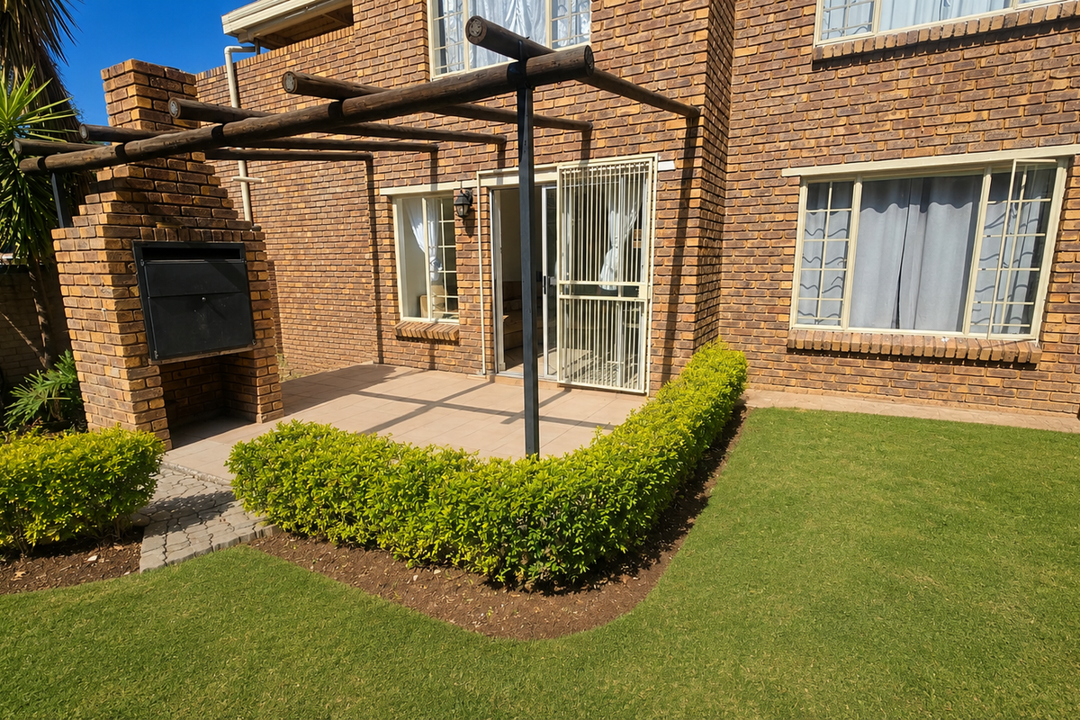 Rear exterior view of the townhouse featuring a brick facade, a paved patio with a built-in braai, a pergola, and a well-maintained garden lawn.