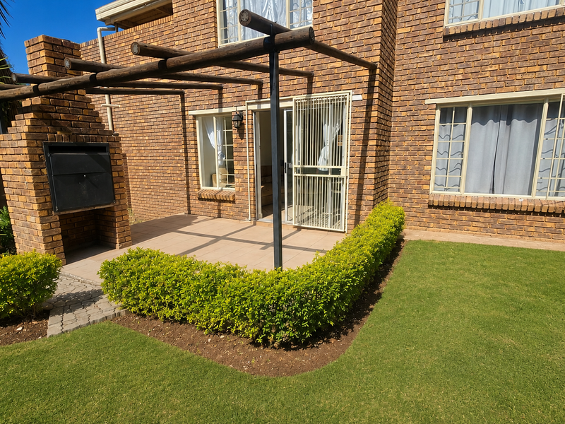Rear exterior view of the townhouse featuring a brick facade, a paved patio with a built-in braai, a pergola, and a well-maintained garden lawn.